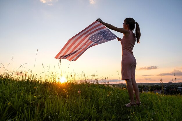 日没時に屋外に立っている米国の国旗とポーズをとって幸せな若い女性の背面図。アメリカ合衆国の独立記念日を祝うポジティブな女性。国際民主主義デー。