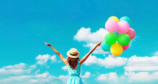 Back view of happy young woman with bunch of colorful balloons on blue sky clouds background