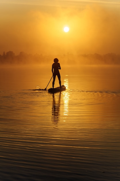 Back view of healthy and active man in silhouette rowing with paddle board on lake.