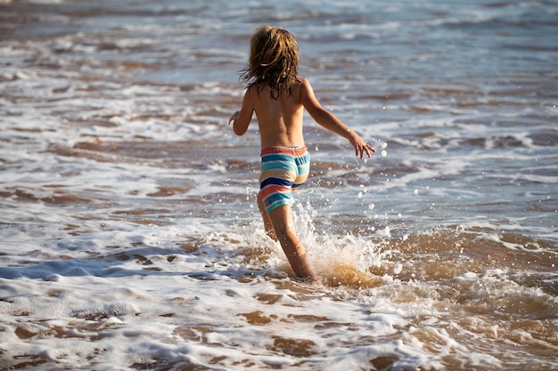 Back view of kid boy have fun on tropical sea beach funny child run with splashes by water pool alon