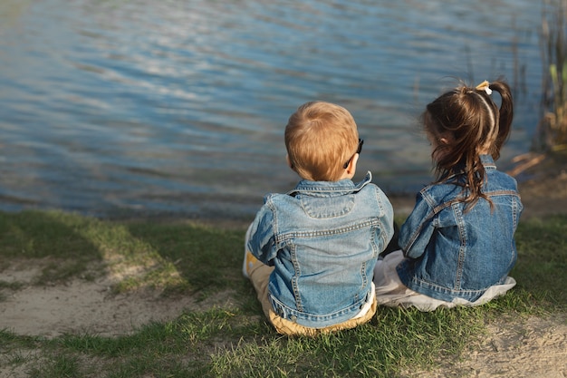 Back view of a little boy and girl sitting on the shore of a pond at sunset