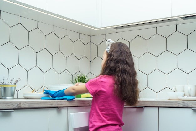 Back view of little preteen girl with loose long dark hair wearing taking off blue rubber gloves standing near table