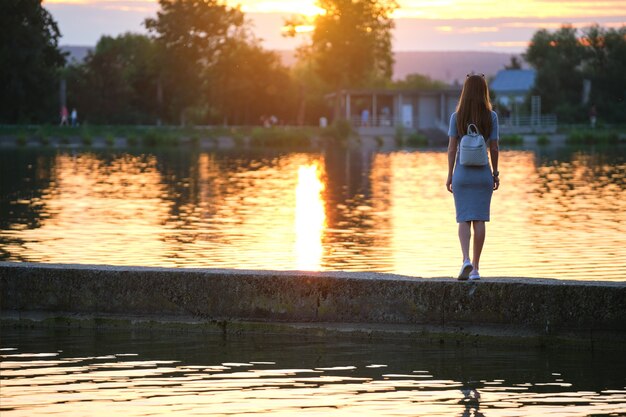 暖かい夜を楽しんで湖岸に一人で立っている孤独な若い女性の背面図。自然の概念の幸福とリラックス。