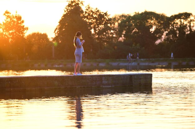 Back view of lonely young woman standing alone on lake shore enjoying warm evening. Wellbeing and relaxing in nature concept.