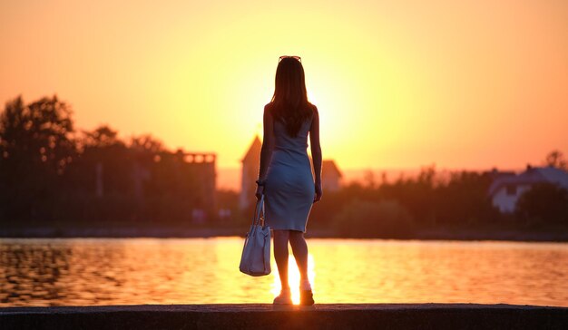 Back view of lonely young woman standing alone on lake shore enjoying warm evening. Wellbeing and relaxing in nature concept.