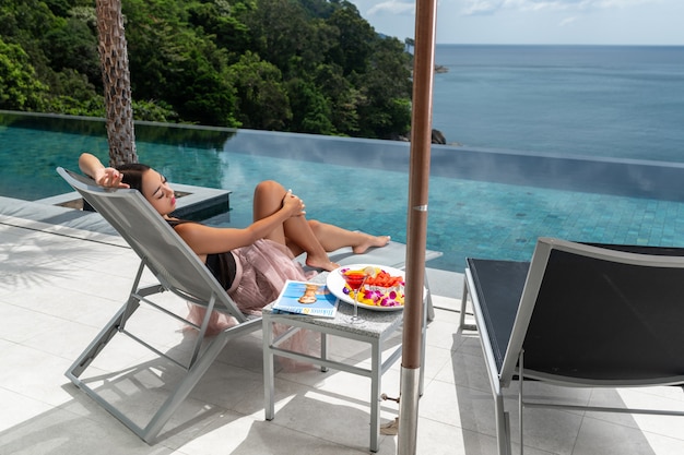 Back view: Luxurious female  lies on a sunbed near the infinity pool. Holidays in tropical countries. Fresh fruits