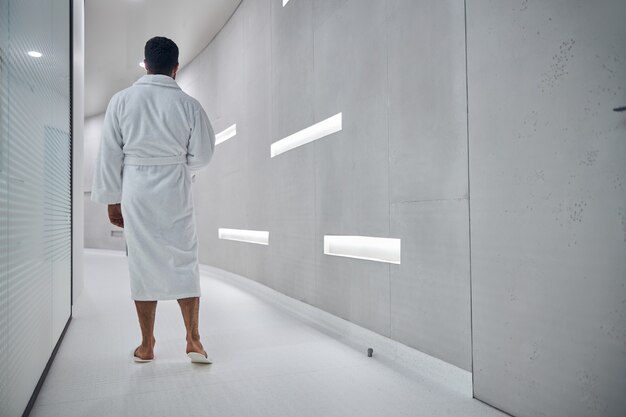 Back view of male client in white bathrobe and slippers spending time in modern wellness clinic