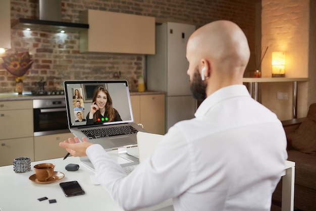 Back view of a male employee in earphones who is working remotely gesturing during a business video conference on a laptop at home. A bald man with a beard an online meeting with his colleagues.