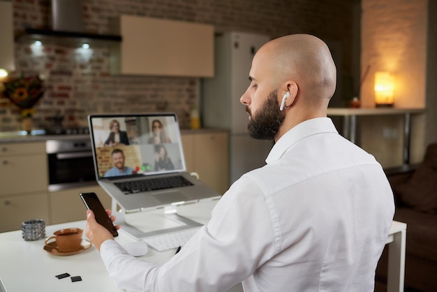 Photo back view of a male employee who is working remotely holding a phone during a business video conference on a laptop at home. a bald man with a beard in earphones on an online meeting with colleagues