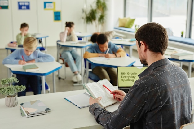 Back view at male teacher sitting at desk in classroom with\
group of children taking test in backgro