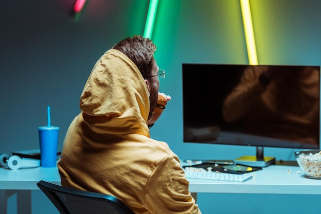 Back view of man eating tasty burger and looking at computer screen with copy space