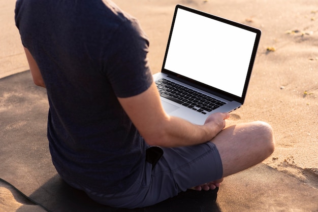 Foto uomo di vista posteriore meditando sulla spiaggia con il suo computer portatile