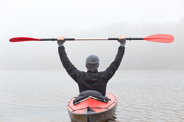 Back view of man rowing kayak across river water in foggy\
morning, raising his paddle up, wearing black jacket and gray\
cap