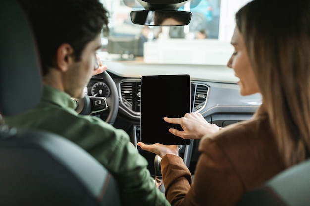 Back view of millennial couple using digital tablet with empty screen sitting in their new car at