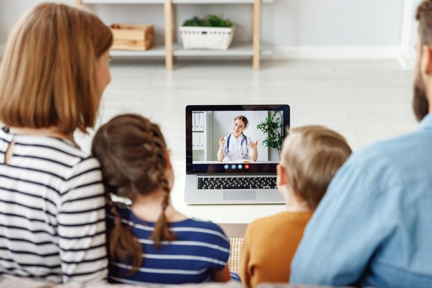 Back view of modern father and children sitting on sofa and
listening to advice of medical practitioner while making video call
to clinic at home