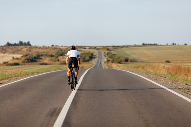 Back view of muscular young man in sport outfit and protective helmet riding bike along countryside