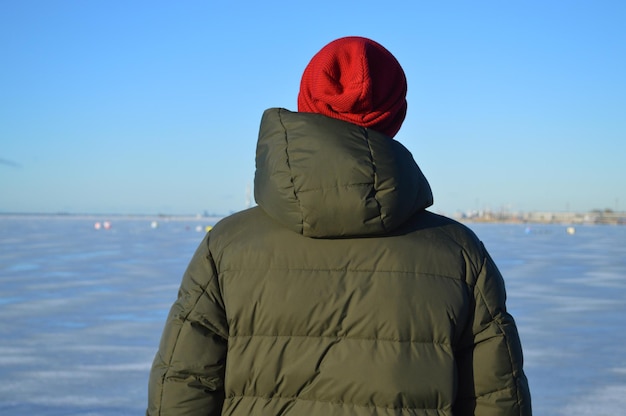 Фото Задний вид человека в зеленой куртке и красной шляпе, глядящего на море