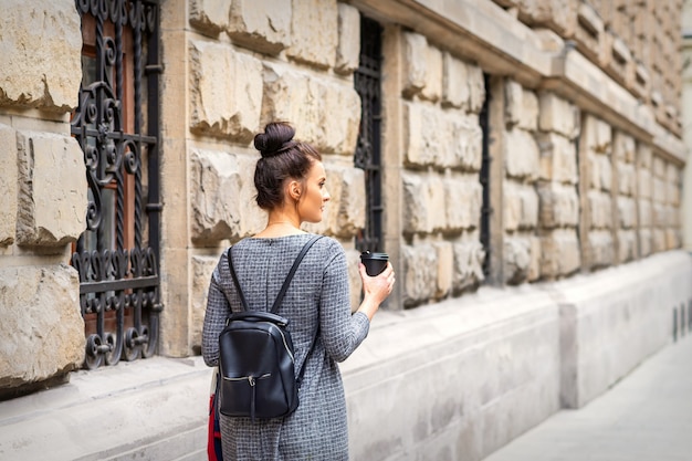 Вид сзади молодой женщины с горячим напитком и рюкзаком гуляет по европейскому городу