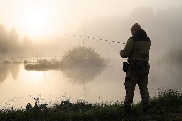 Вид сзади анонимного рыбака, стоящего на берегу пруда и ловящего рыбу ранним туманным утром
