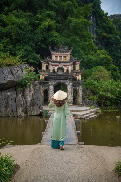 Фото Задний вид девушки, одетой в ао дай перед открытым парком с озером и каменным мостом вход в древний комплекс пагоды бич донг нинь бинь вьетнамское туристическое направление