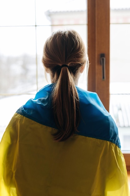 写真 家で窓の外を見ている旗に包まれた金髪の女性サポートウクライナの背面図ウクライナの概念での戦争