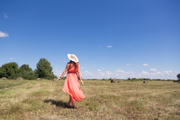 写真 赤い夏のドレスと黄色の農場の牧草地に立っている麦わら帽子の若い幸せな女性の背面図