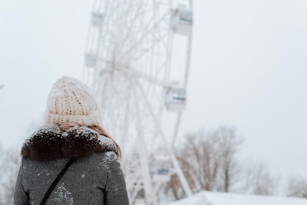 屋外の公園で冬の散歩中に観覧車の魅力を見ている雪に覆われたコートとニットの白い帽子の若い女性の背面図。スペースをコピーします。
