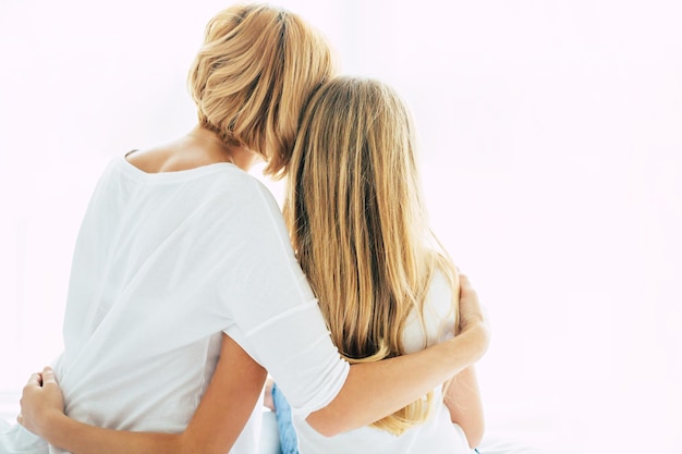 Back view photo of beautiful young mother with her little daughter sitting on the bed on white window backround