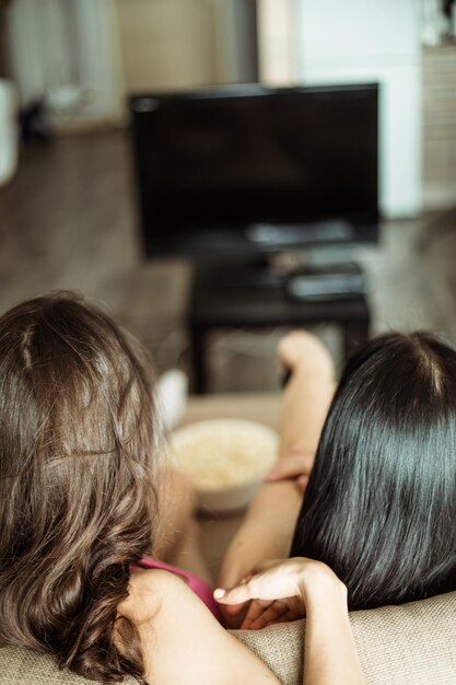 Фотография сзади двух подруг, которые сидят на диване и смотрят телевизор