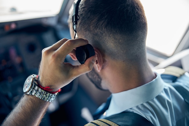 Vista posteriore ritratto del pilota barbuto con l'orologio a portata di mano mentre si tocca le cuffie nella cabina di pilotaggio dell'aereo