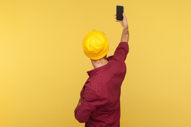 Back view portrait of hipster guy in beanie hat and checkered\
shirt talking video call taking selfie on mobile phone holding\
smartphone up with mock up blank display indoor studio shot\
isolated