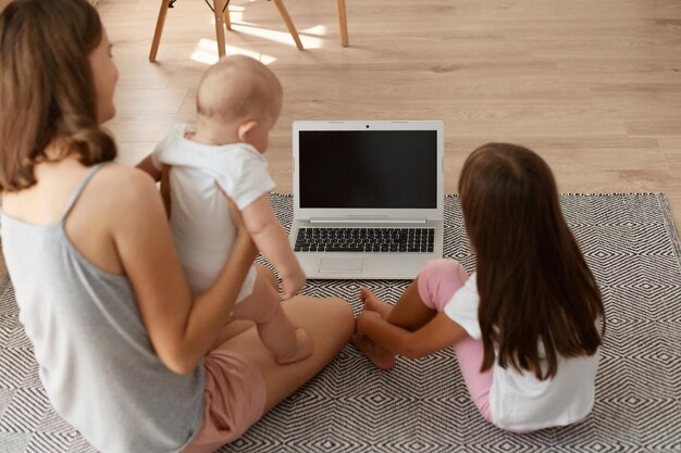 Back view portrait of woman wearing casual clothing sitting on floor with her little children and looking at display of notebook, blank screen with mockup for commercial advertisement.