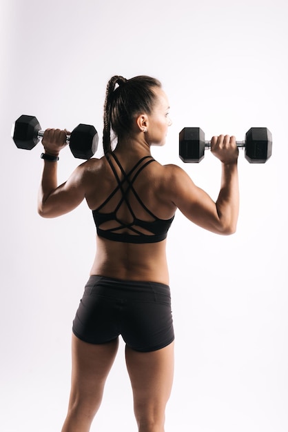 Back view portrait of young muscular woman bodybuilder with perfect athletic body in sports black bra Sporty muscular girl is lifting dumbbells at shoulder level on isolated white background