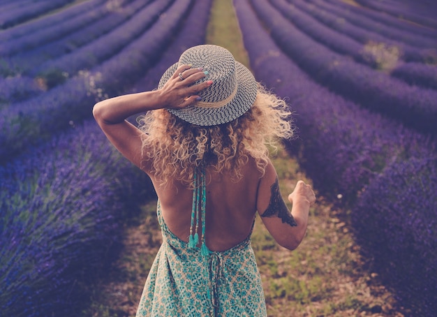 보헤미안 우아한 파란색 드레스를 입은 예쁜 아기의 뒷모습은 여행 스타일 모자와 금발의 곱슬곱슬한 긴 머리를 입고 라벤더 밭에서 산책 - 개념 또는 야외 자연 모험 생활 방식의 자유로운 여성