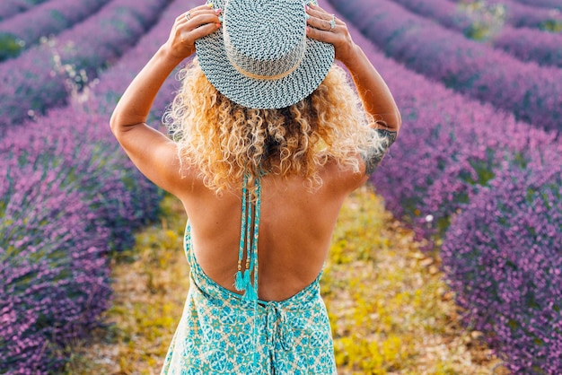 Back view of pretty curly blonde woman holding trendy hair and admiring a scenic lavender field Travel female people lifestyle Elegant rear of female enjoying nature and scenery destination