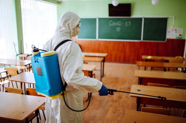 Back view of a professional sanitary worker disinfecting the classroom before the academic year starts. A man wearing protective suit cleans up the auditorium from coronavirus Covid-19. Health care.