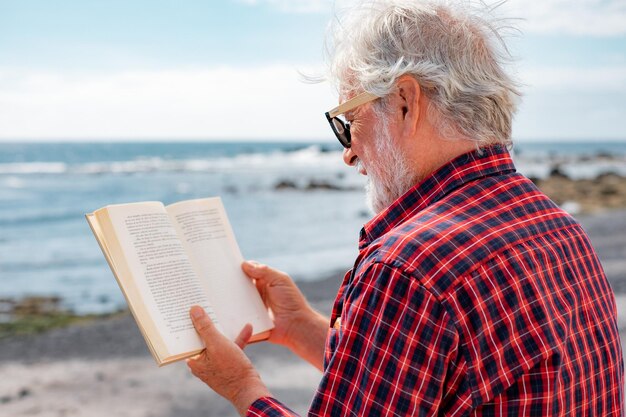 Back view of senior bearded man reading a book in front to the sea Elderly relaxed pensioner