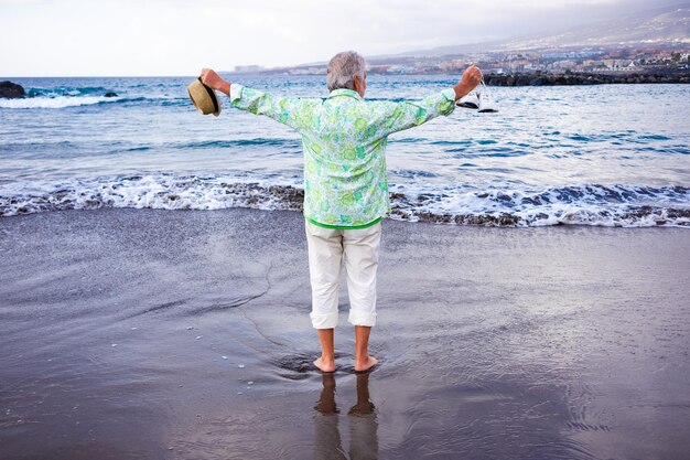 Back view of senior grayhaired man standing on the beach barefoot holding shoes and hat in hands enjoying vacation looking at horizon over water Concept of relaxed and free retiree