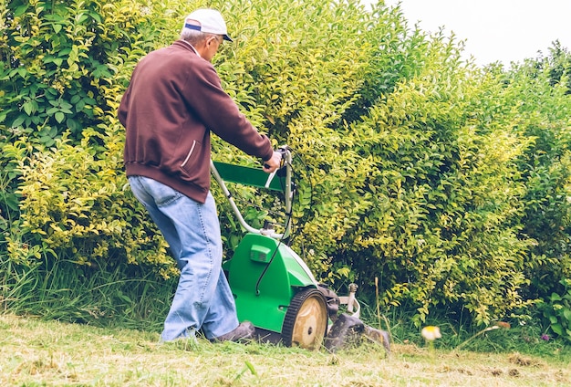 Vista posteriore di un uomo anziano con cappuccio che falcia il prato con una macchina tosaerba in un campo