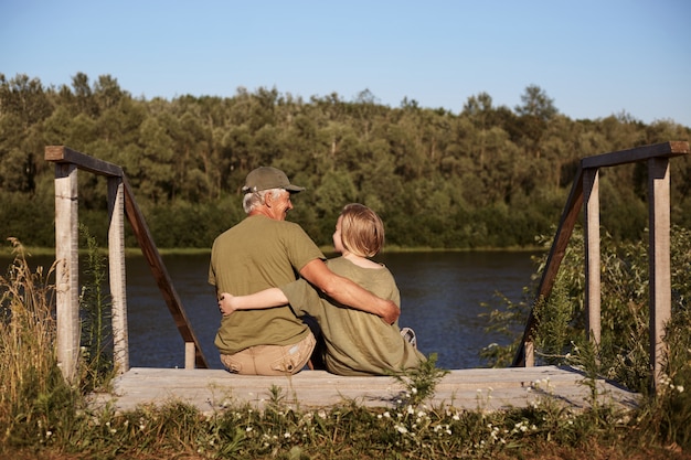 Back view of senior man with his grandson sitting at wooden setting near river outdoors