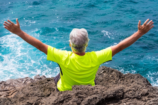 Photo back view of senior white-haired man in outdoor excursion at sea, sitting on the rocks looking at horizon with arms outstretched