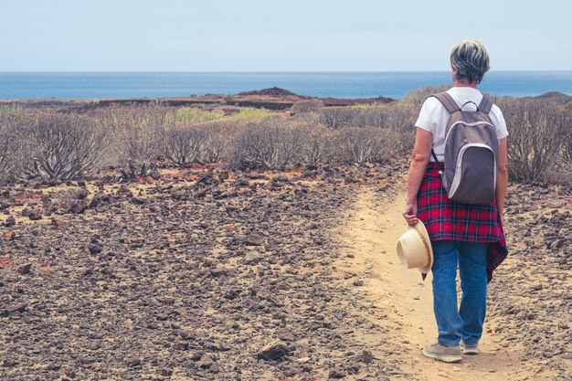 Back view of senior woman with backpack in outdoor trekking, looking at horizon over sea.