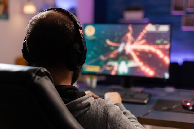 Back view shot of concentrate gamer streaming online videogames on computer using wireless controller. Player man with headphones playing games in room with neon light and professional equipment