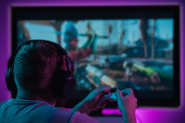 Back view shot of professional gamer playing online video game on his gameset. Room lit in neon light and retro style. Using headphones to talk with the team. Entertainment, fun concept.