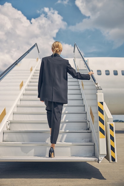 Вид сзади стройной блондинки в брючном костюме, поднимающейся по ступенькам самолета