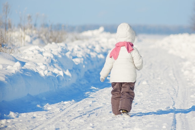 Back view of small female kid wears warm winter clothes, strolls during frosty winter weather, likes to play outdoors