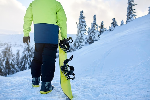 Back view of snowboarder climbing with his board on the mount for backcountry freeride session in the forest Man with snowboard walking at ski resort Rider lime fashionable outfit