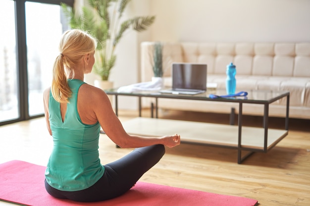 Back view of sportive mature blonde woman sitting in lotus position on the floor and doing