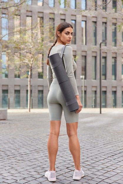 Photo back view of sporty muscular brunette woman dressed in sportsclothes carries rolled karemat dressed in sportsclothes and sneakers turns behind returs after aerobics or gymnastics stands outdoors