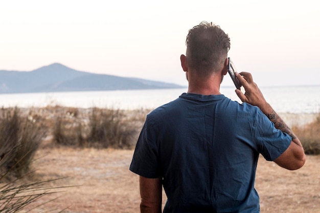 Вид сзади на стоящего человека, использующего мобильный телефон и звонящего по телефону на открытом воздухе на фоне живописного места назначения с океаном и островом. Люди, туристы и мобильные телефоны, подключение к Wi-Fi.